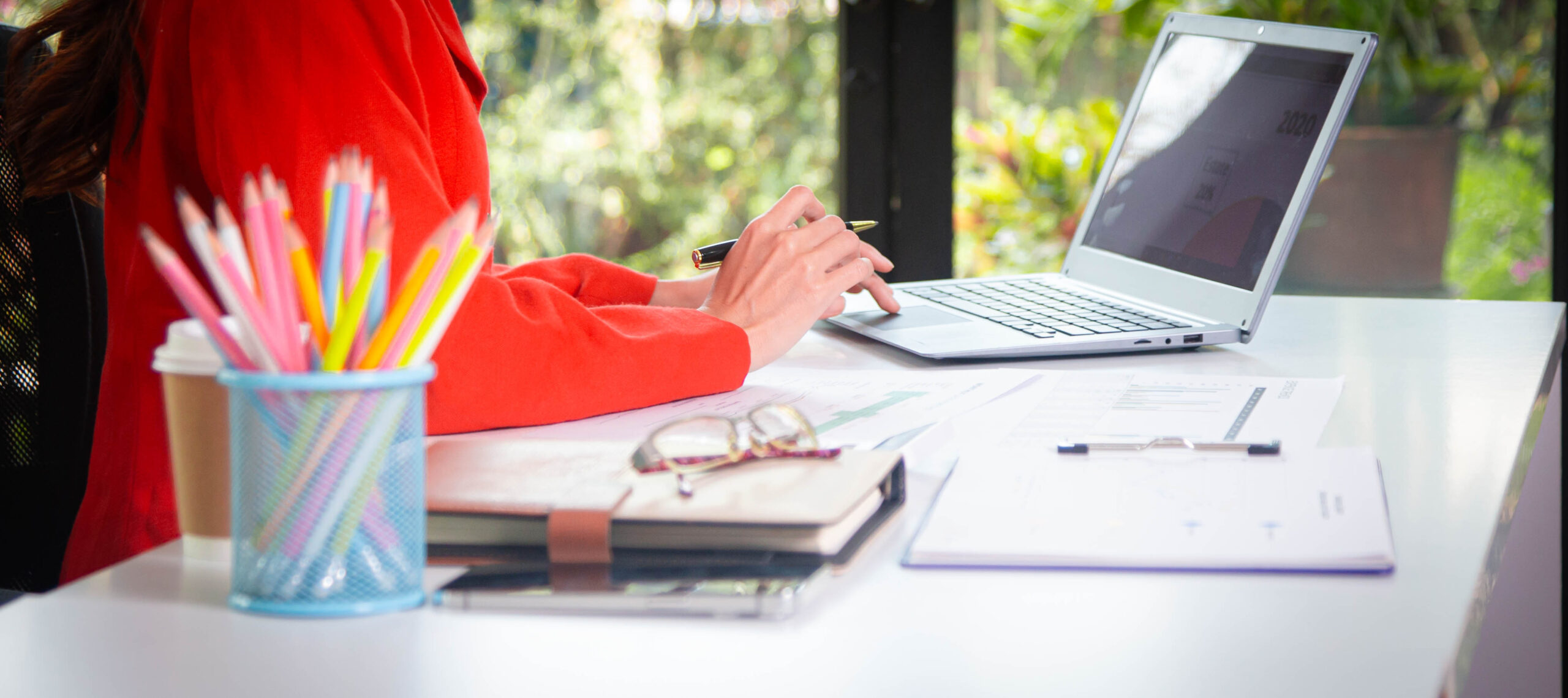 femme avec un veston rouge assise à un bureau de travail devant un ordinateur portable avec vue sur la nature